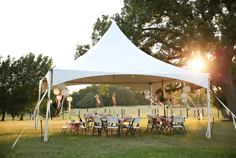 a square marquee event tent setup in a field with plastic folding chairs and round tables underneath it.